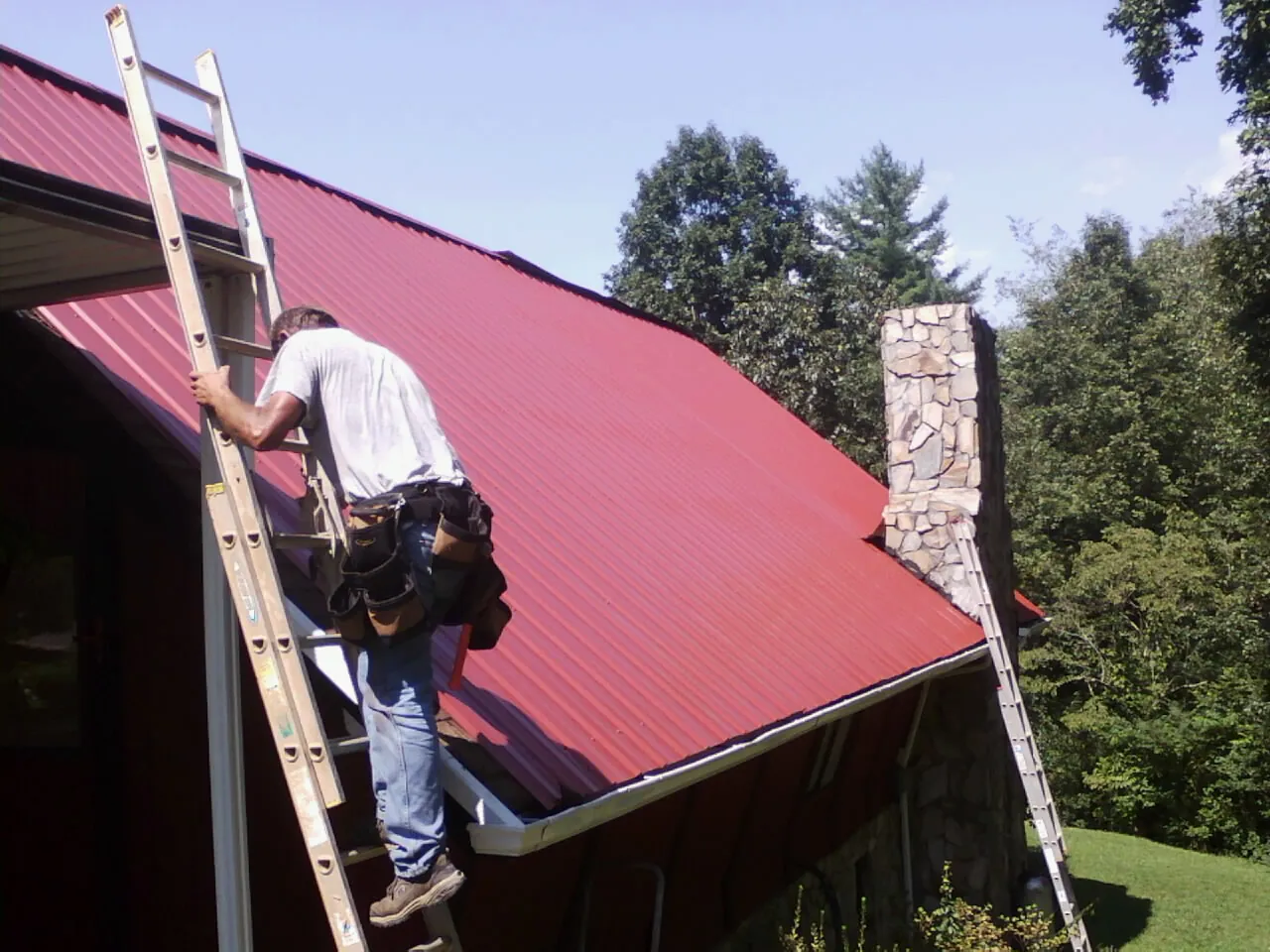 Roofer on a ladder beside a red metal roof and stone chimney — Soffit & Fascia Repair by Roofing Brothers in Cook
